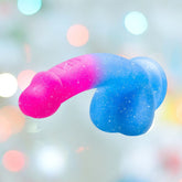 The Avant Chasing Sunsets Dildo - Mermaid by Avant features a sparkly pink-to-blue gradient and a suction cup, set against a soft pastel background with blurred circular light spots.
